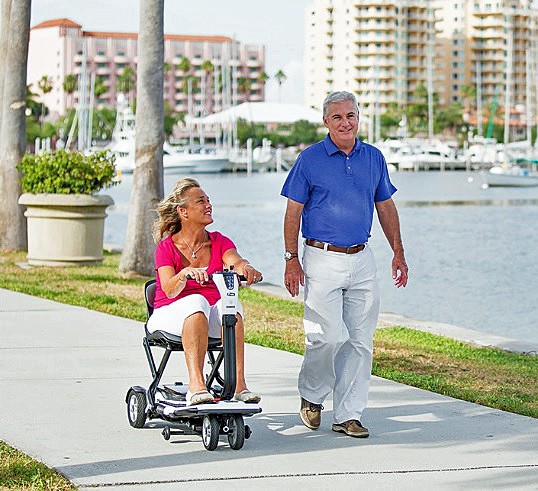 Go-Go Folding 4-Wheel Travel Scooter - Happy Casual Couple Walking and Riding by the Coast on a Sunny Day